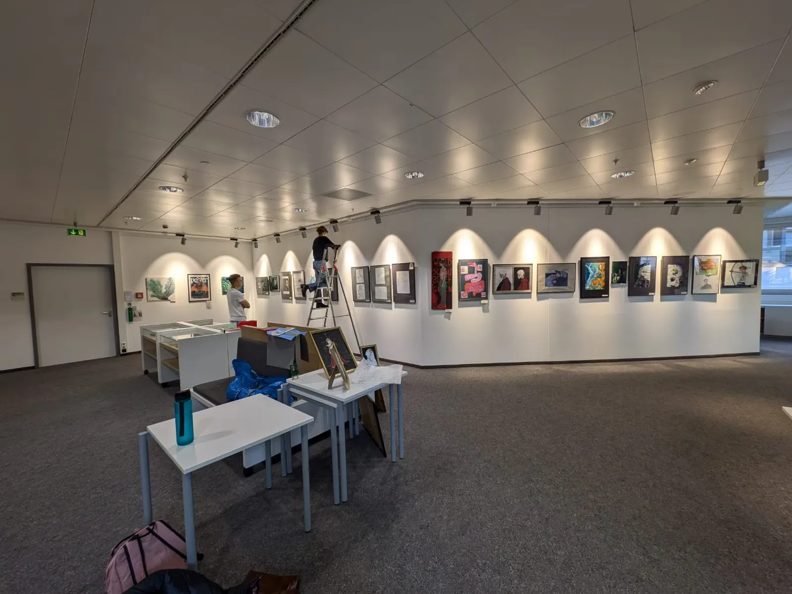 Blick auf eine Wand der Jubiläums-Ausstellung des MWG-Kunstclubs. Eine Person steht auf einer Leiter und hängt ein Bild auf.